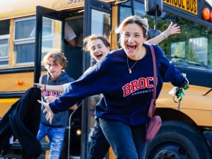 Three students, one smiling broadly in a Brock University sweatshirt, step off a yellow school bus, as they arrive for an off-campus event.