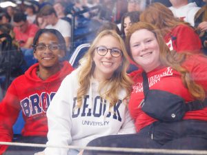 Three people in Brock University hoodies and T-shirts smile while sitting in an arena.