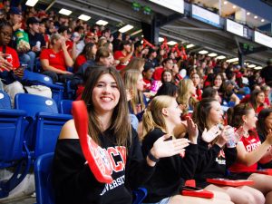 A woman with a large foam finger on her hand cheers in a crowd full of people at an arena.
