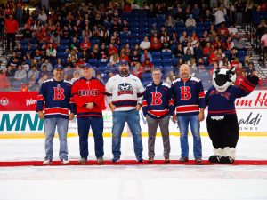 Five men and Boomer the Badger mascot stand on the ice in an arena with fans in the background in the stadium seats.