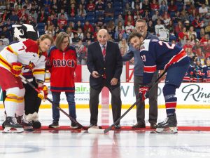 A man in a suit drops a hockey puck at centre ice while two hockey captains face off in a ceremonial puck drop. They are joined by two other inidviduals and Brock's mascot, Boomer the Badger.