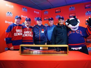 A group of five men join Boomer the Badger mascot, standing behind the Steel Blade Classic trophy, a large, silver sword encased in a wooden box.
