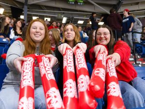 Three women hold Brock Badgers thunder sticks — large inflatable sticks meant to be hit together during a sporting event — while sitting in a busy arena.