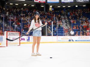 A woman in a Brock University sweatshirt holds a hockey stick while standing on the ice in an arena, preparing to hit a hockey puck.