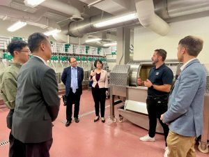 A group of people listen to a man speak in a wine-making facility.