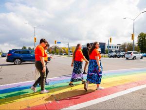 A group of people wearing orange shirts and ribbon skirts walk together across a rainbow-painted crosswalk at a busy intersection, participating in a community event.