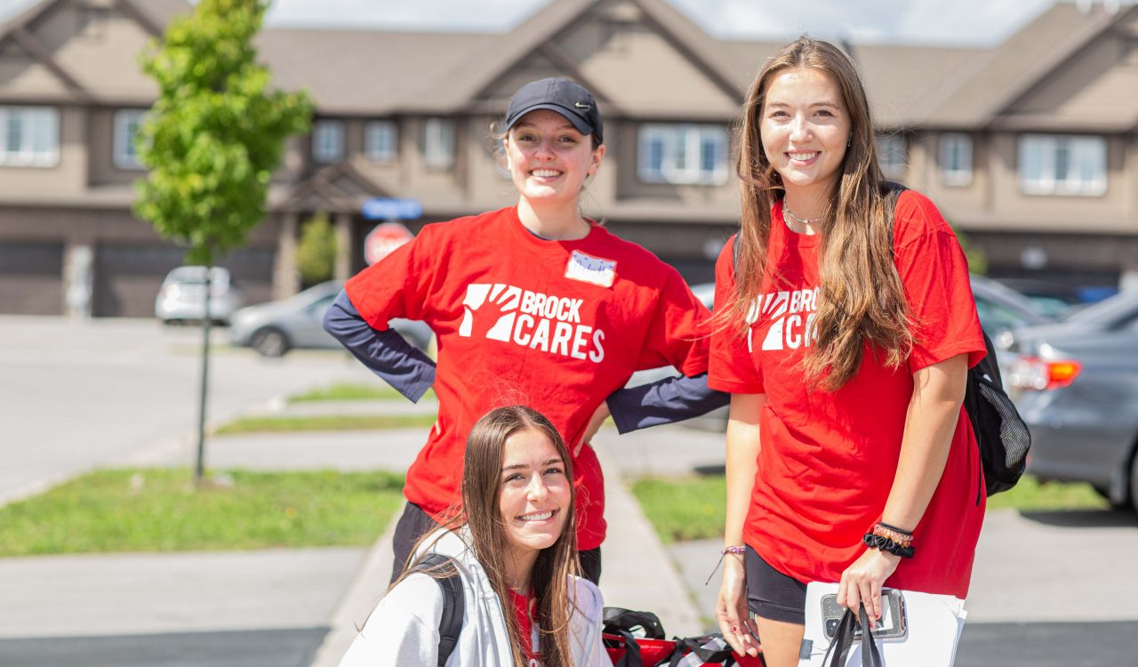 Three university students in red shirts smile outside a row of houses.