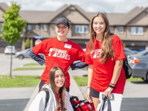 Three university students in red shirts smile outside a row of houses.