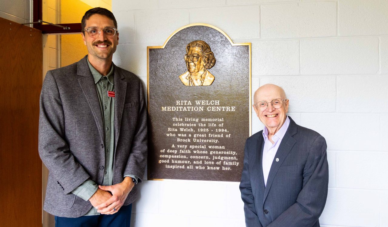Two men stand in front of a plaque that says Rita Welch Meditation Centre.