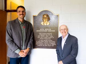 Two men stand in front of a plaque that says Rita Welch Meditation Centre.