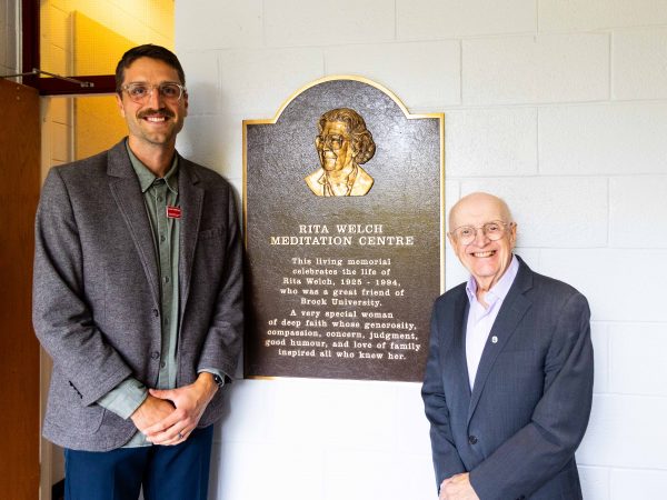 Two men stand in front of a plaque that says Rita Welch Meditation Centre.