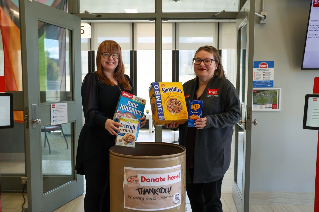 Two women hold up grocery items beside a bin for a food drive.