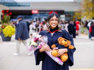 A smiling graduate holds flowers and a teddy bear outside the ceremony venue.