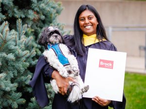 A graduate poses with her dog and degree folder in front of a pine tree.