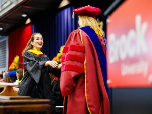 A graduate enthusiastically shakes hands with a faculty member on stage.