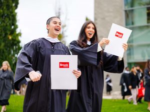Two graduates cheerfully hold their Brock University degree folders outdoors.