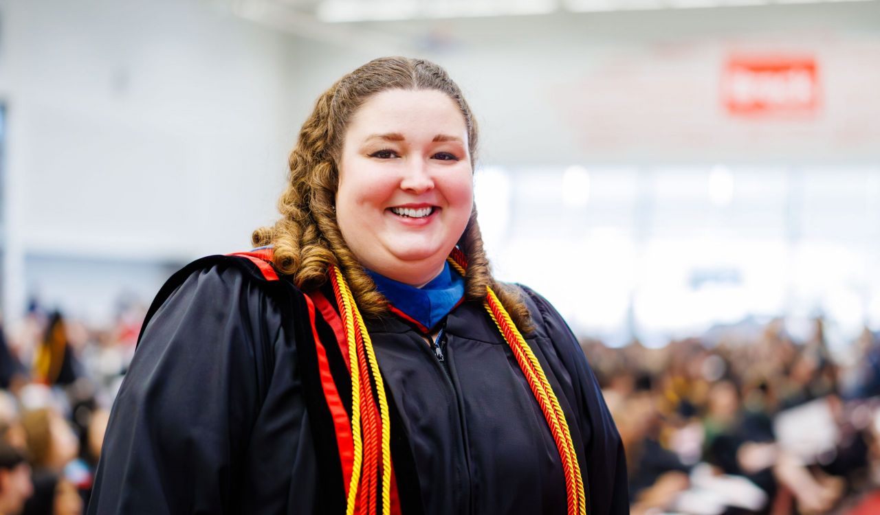 Woman wearing a black graduation robe and cords stands and smiles in front of a crowd of people during Brock University's 118th Convocation.