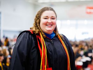 Woman wearing a black graduation robe and cords stands and smiles in front of a crowd of people during Brock University's 118th Convocation.