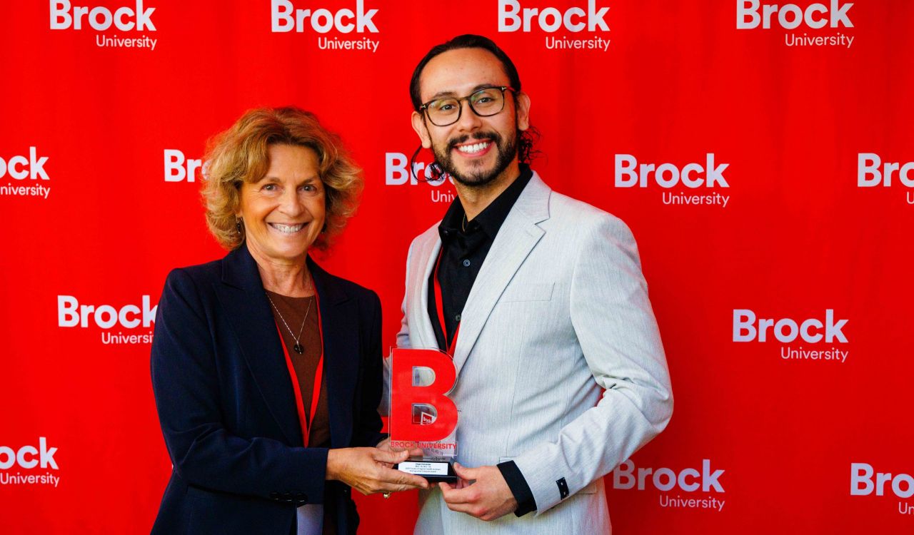 A man and a woman pose with an award trophy in front of a red backdrop.