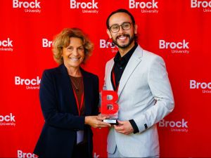 A man and a woman pose with an award trophy in front of a red backdrop.