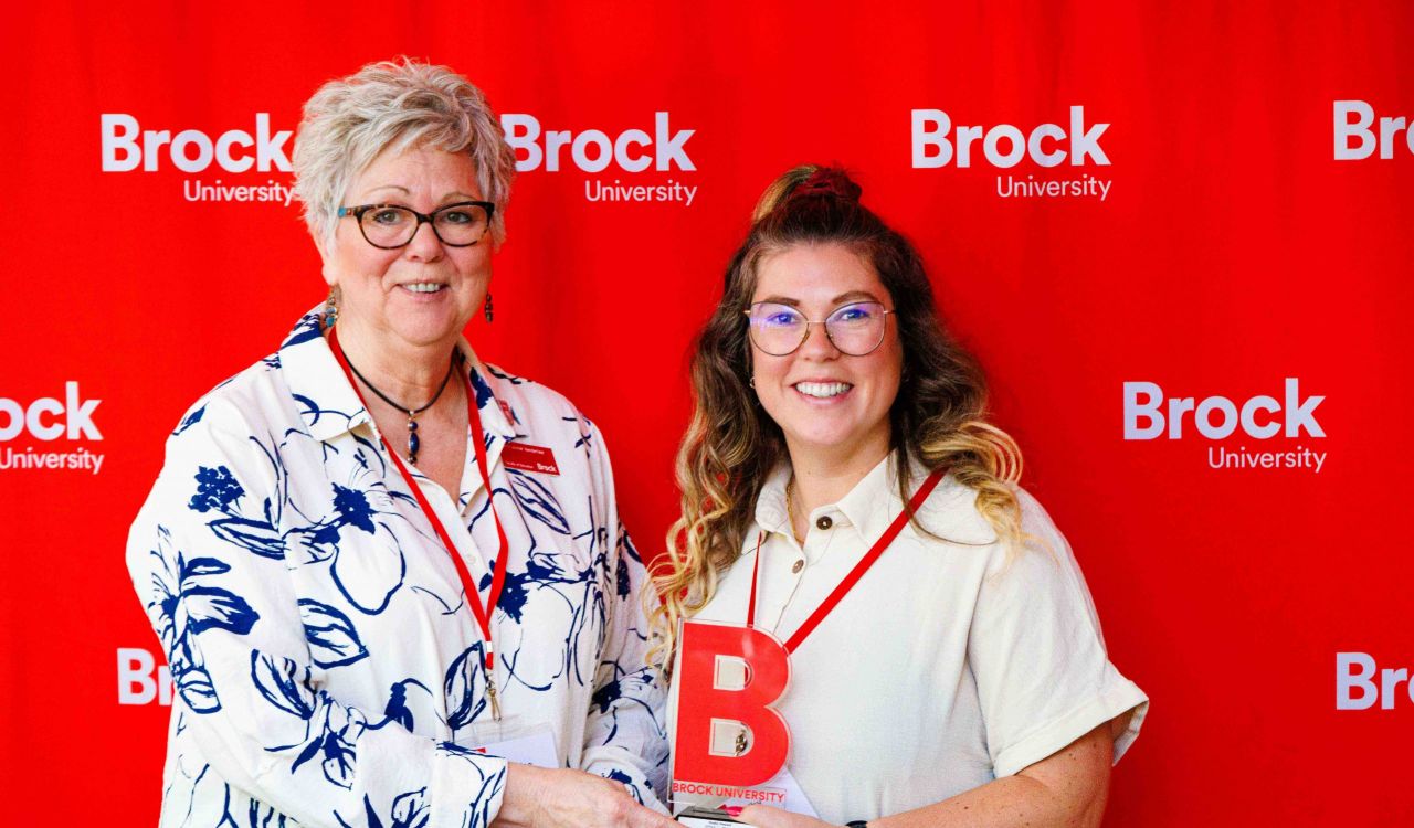 Two women stand side by side smiling holding an award in front of a red Brock University backdrop.