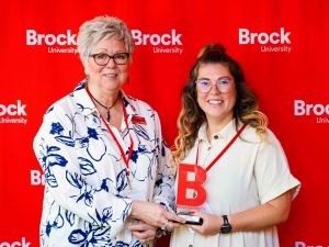 Two women stand side by side smiling holding an award in front of a red Brock University backdrop.