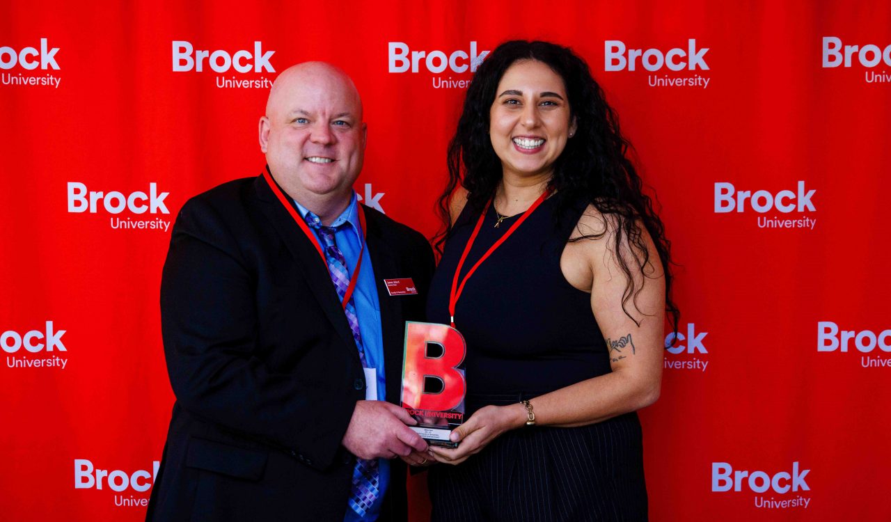 A man stands beside a woman handing her an award in front of a Brock University red background.