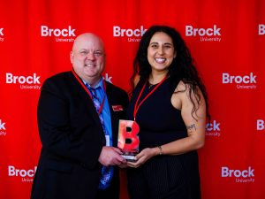 A man stands beside a woman handing her an award in front of a Brock University red background.
