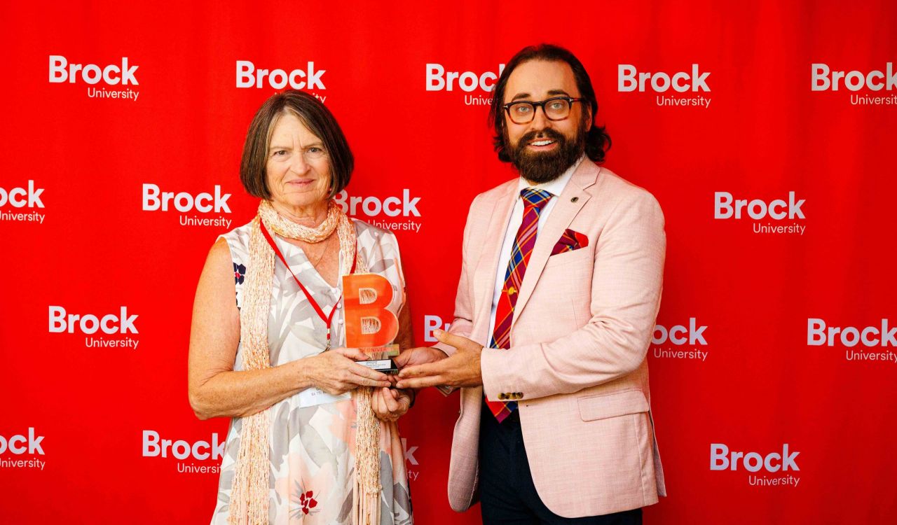 Two people pose in front of a red backdrop with a trophy.