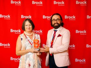 Two people pose in front of a red backdrop with a trophy.