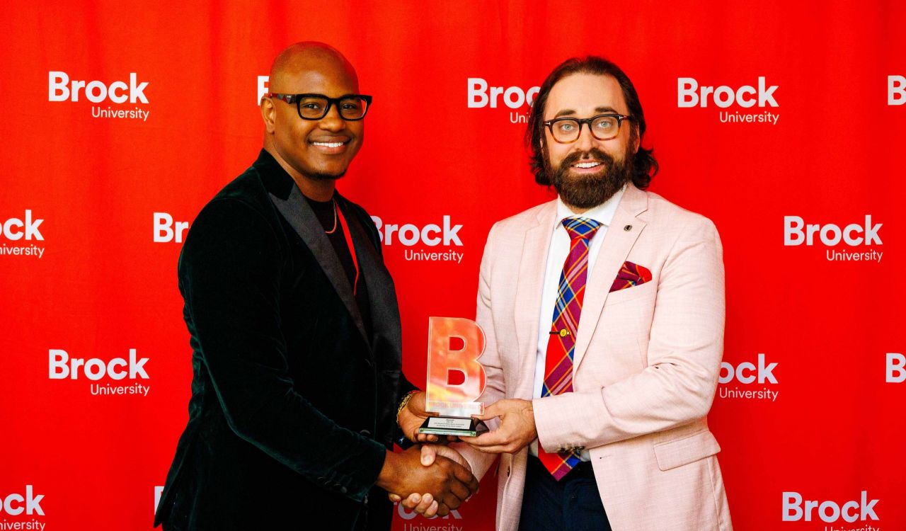 Two men pose in front of a red screen with an award.