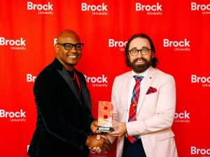 Two men pose in front of a red screen with an award.