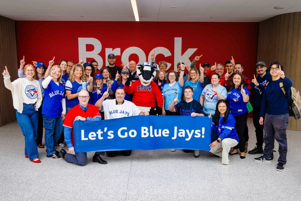 A large group of people pose behind a "Let's go Blue Jays!" banner. Many are wearing Toronto Blue Jays shirts or hats.