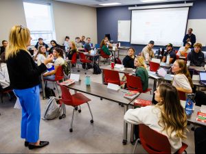 A classroom full of students seated at tables with laptops and notebooks listens attentively to a woman standing at the front of the room.