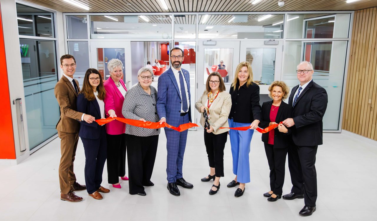 A group of nine people stand side by side indoors for a ribbon-cutting ceremony. They hold a long red ribbon stretched across the entrance of a bright, modern space with glass doors and wood-paneled ceiling. One woman in the center holds large ceremonial scissors, smiling as she prepares to cut the ribbon while others on both sides smile toward the camera.