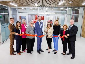 A group of nine people stand side by side indoors for a ribbon-cutting ceremony. They hold a long red ribbon stretched across the entrance of a bright, modern space with glass doors and wood-paneled ceiling. One woman in the center holds large ceremonial scissors, smiling as she prepares to cut the ribbon while others on both sides smile toward the camera.