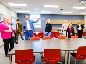 A group of visitors tour a modern classroom with red chairs and wooden tables.