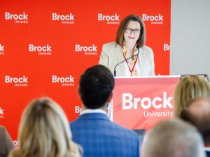 A speaker addresses an audience from a podium at a Brock University event, with the university’s red logo backdrop displayed behind them.