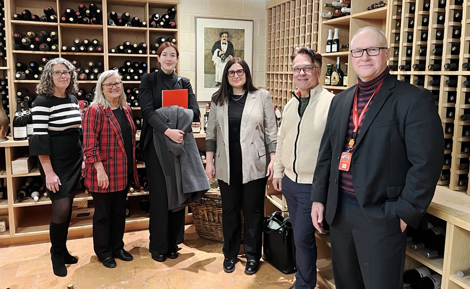 Six people stand together in a large wine cellar.