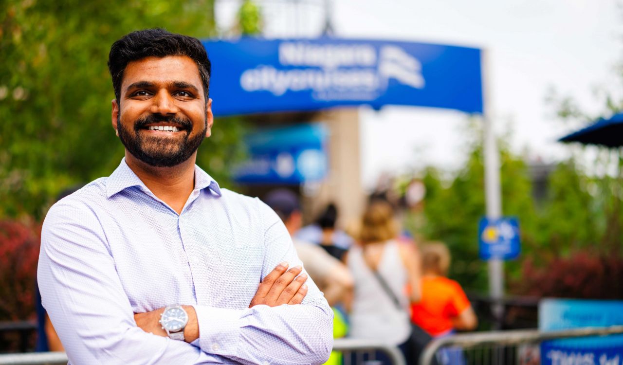 A smiling man wearing a dress shirt stands confidently with his arms crossed in front of the Niagara City Cruises entrance. People are gathered in the background on a sunny day.