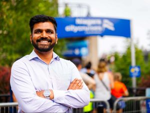 A smiling man wearing a dress shirt stands confidently with his arms crossed in front of the Niagara City Cruises entrance. People are gathered in the background on a sunny day.