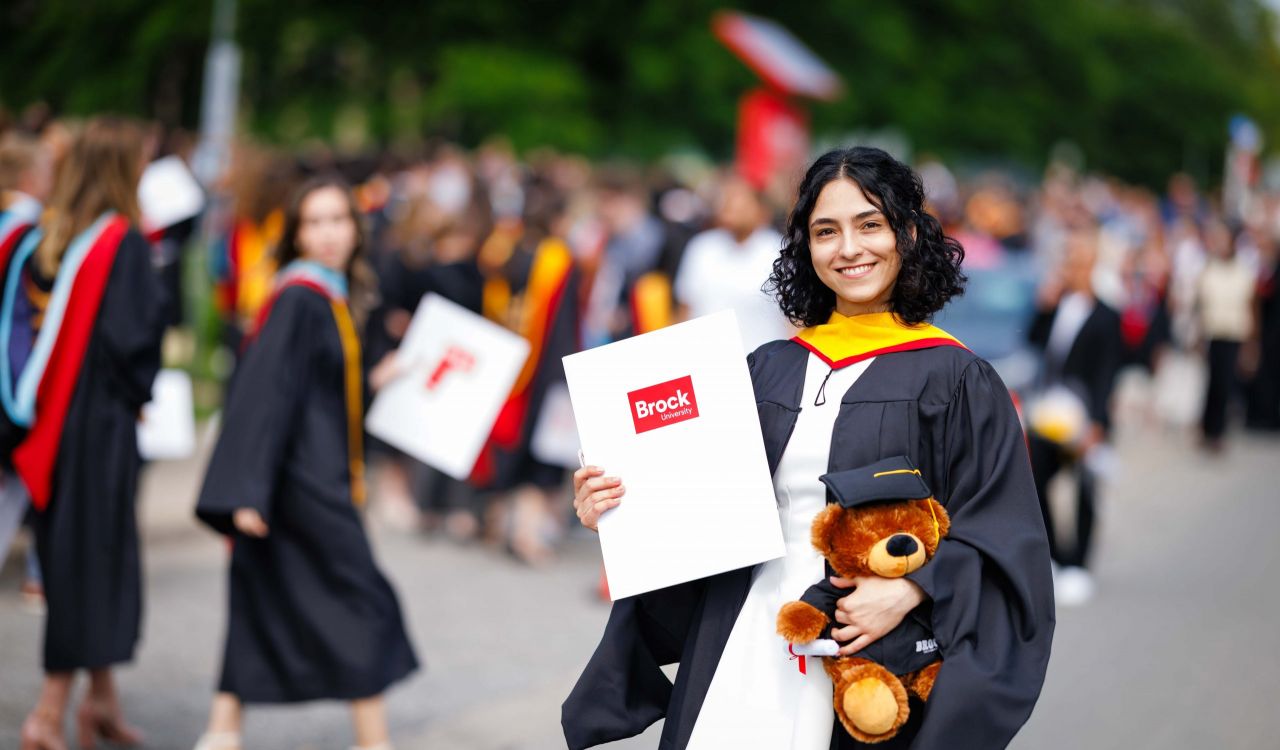 A young woman in a university graduation gown poses with a diploma outside after a graduation ceremony.