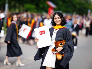 A young woman in a university graduation gown poses with a diploma outside after a graduation ceremony.