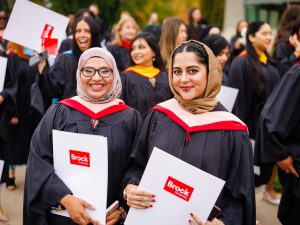 Two women wearing academic regalia smile and stand side-by-side holding degrees during Brock University's 118th Convocation.