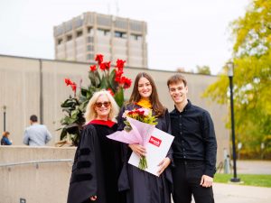 Three people stand side-by-side in academic regalia and hold flowers and degrees during Brock University's 118th Convocation.