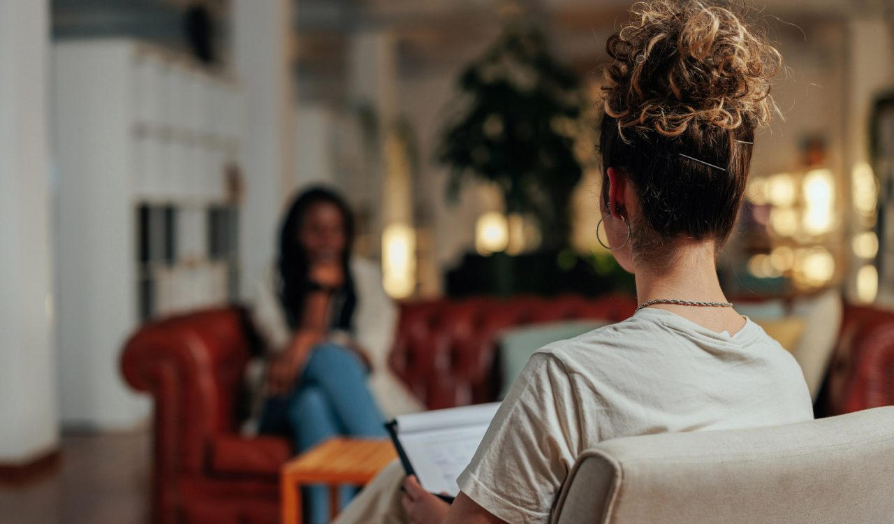 Close-up view of the back of a therapist's head, with a patient, who is out of focus, sitting across from her on a couch.