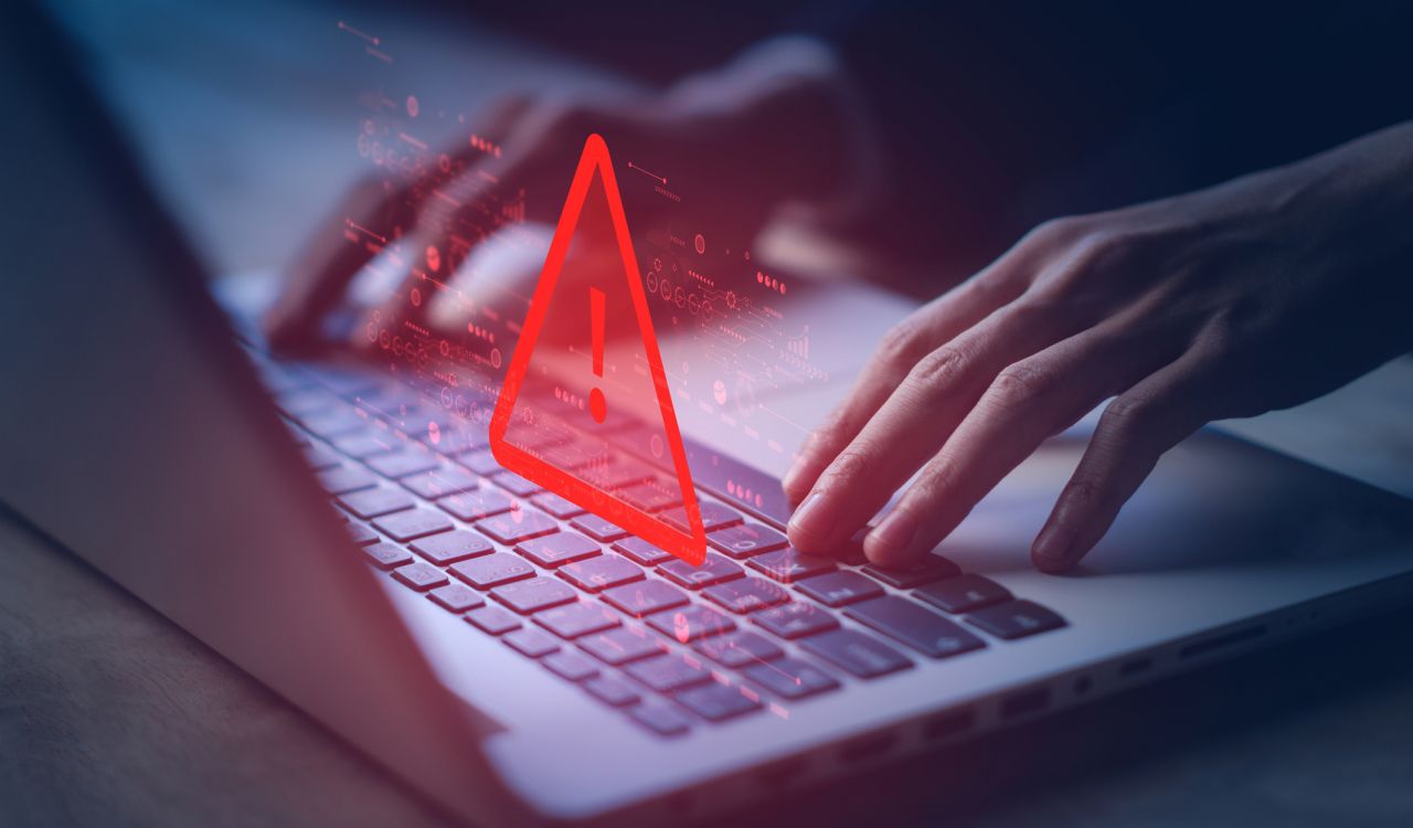 A warning sign hovers over hands typing on a laptop.