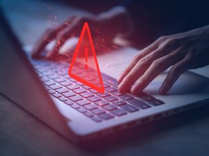 A warning sign hovers over hands typing on a laptop.