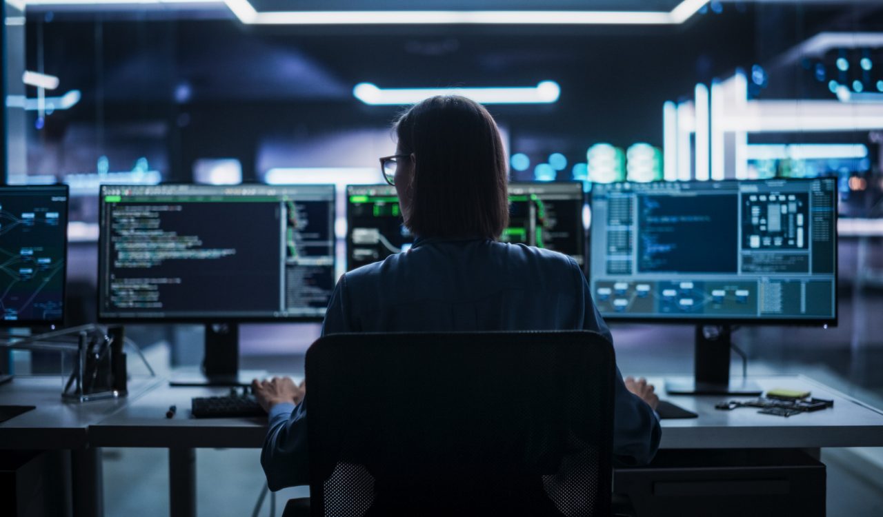 Silhouette of a woman sitting in front of several computer screens in a darkened room.