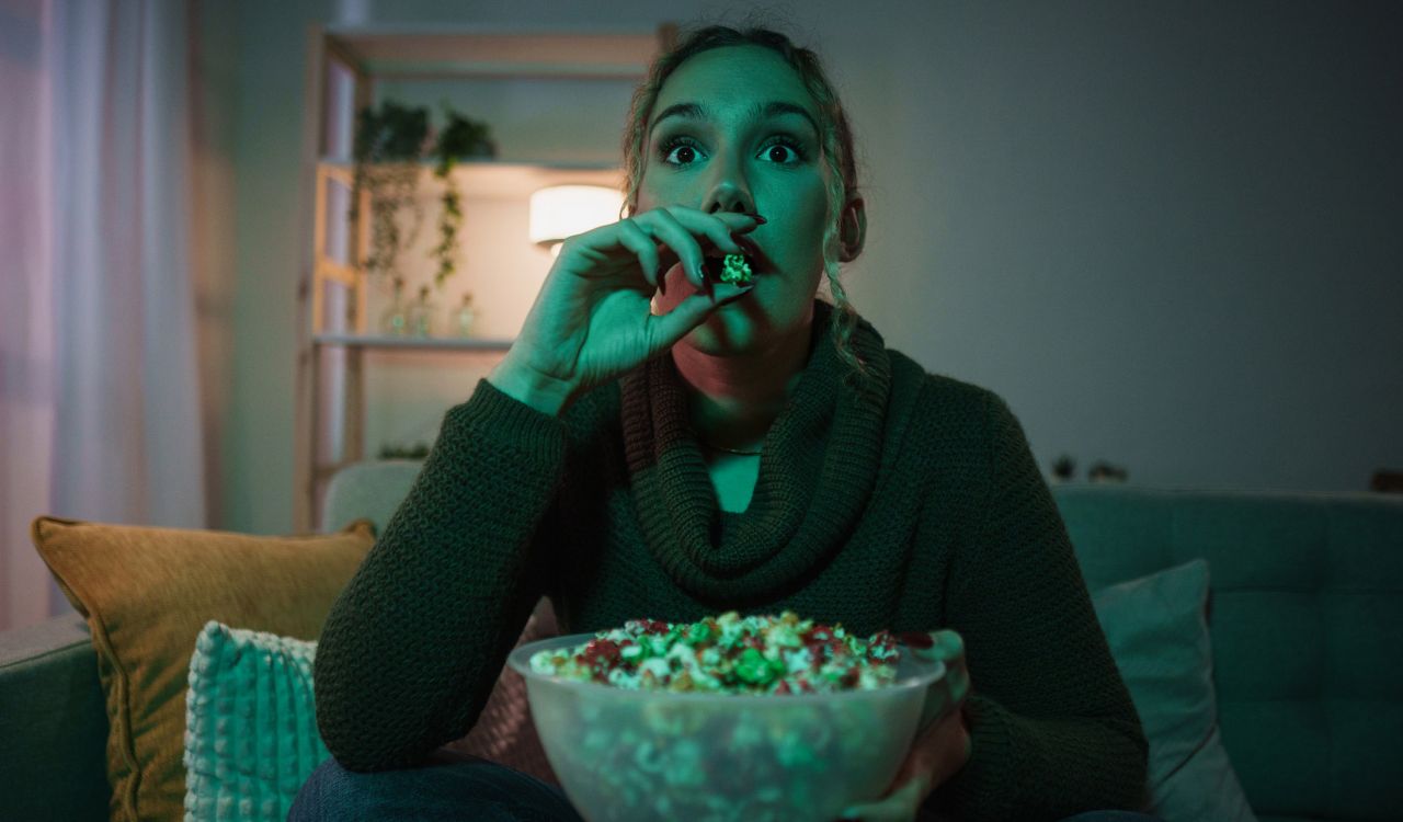A woman, who looks startled, eats popcorn while watching television.
